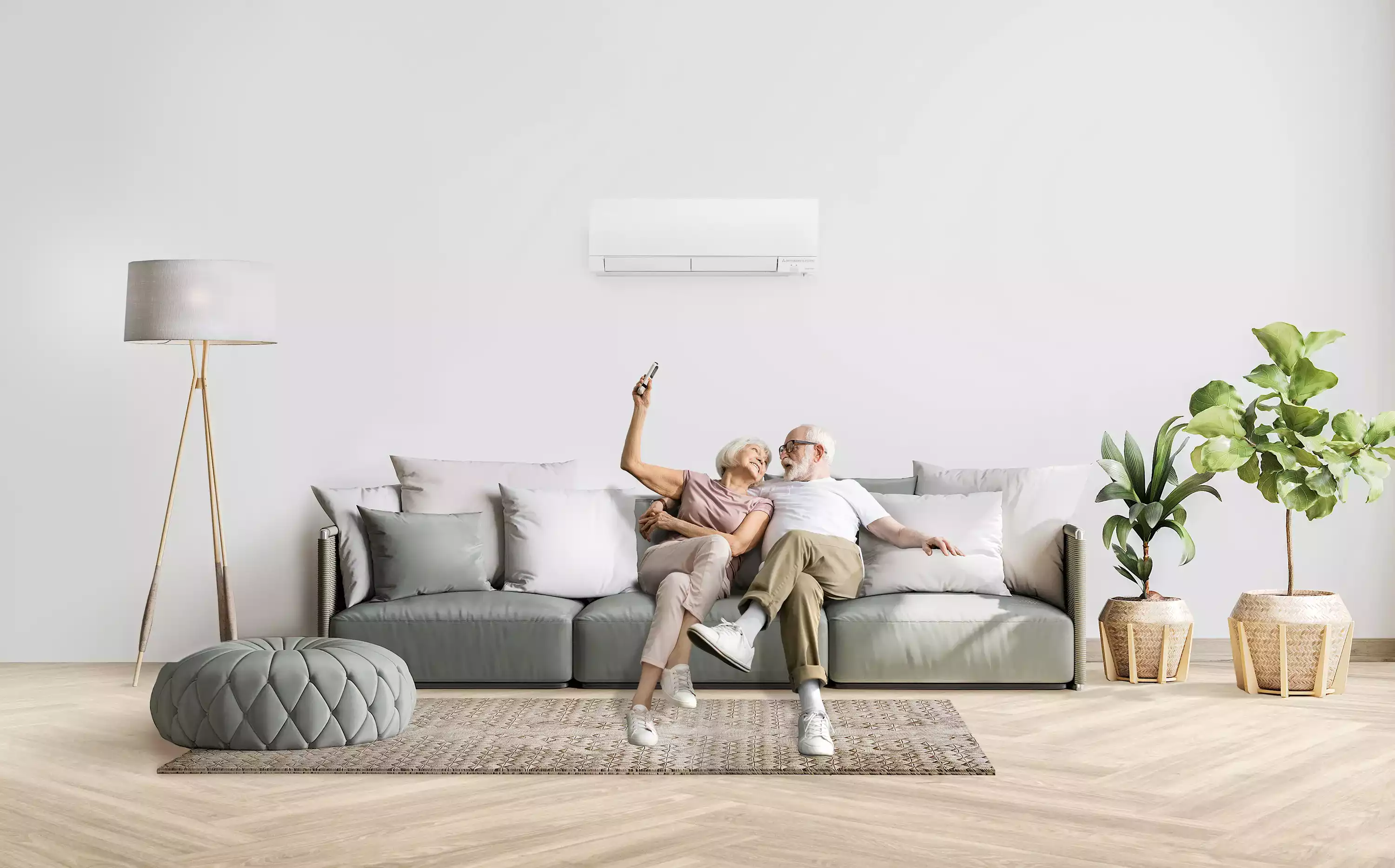 Couple âgé assis sur un canapé, souriant et tenant une télécommande, sous un climatiseur mural dans un salon moderne.